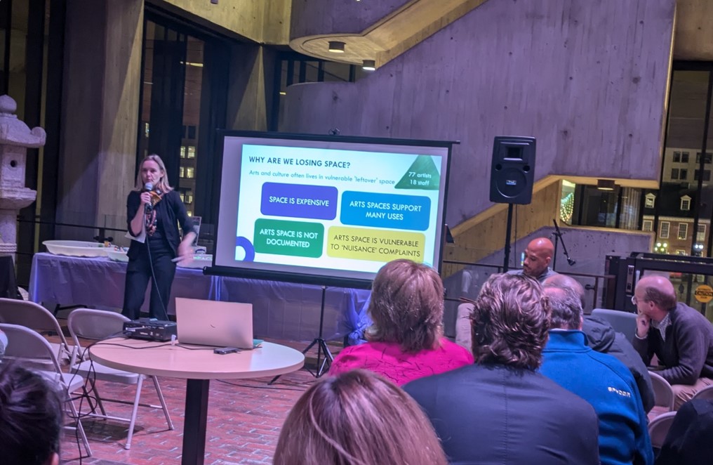 A woman stands to the right of a screen displaying a colorful slide titled "Why are we losing space" with four word bubbles showing the text "Space is Expensive," "Arts spaces support many uses," "Arts space is not documented," and "Arts space is vulnerable to nuisance complains." To the right sit two men. In the foreground are the backs of audience members and a table holding a laptop and projector.