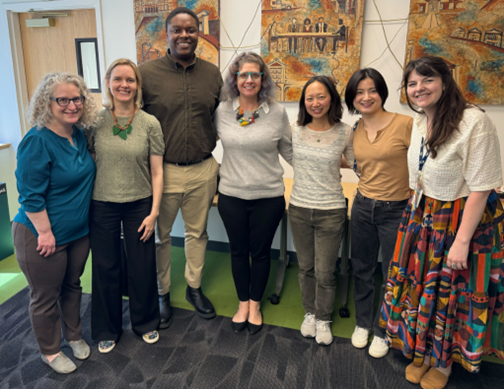 Group photo of seven people standing and smiling. Pictured are members of the MAPC Arts and Culture team, Jenn Chang, and Dee Schneidman—Senior Program Director for the Creative Economy at the New England Foundation for the Arts. They are gathered in a brightly lit room with colorful artwork on the wall behind them.