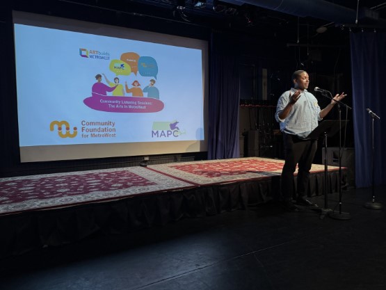 Lafayette Cruise, Arts and Culture Planner II at MAPC, stands on stage speaking at a microphone during a Community Listening Session. A large screen behind him displays logos for the Community Foundation for MetroWest, MAPC, and Arts & Culture MetroWest, along with the event title: "Community Listening Sessions: The Arts in MetroWest."