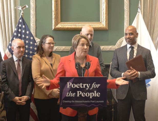Governor Maura Healey speaks at a podium with a sign reading “Poetry for the People: Creating the Massachusetts Poet Laureate.” She is wearing a red blazer and gesturing with both hands. Behind her, four individuals stand, including Lieutenant Governor Kim Driscoll and Mass Cultural Council Executive Director Michael J. Bobbitt, along with an American flag and a Massachusetts state flag in a room with green walls and a gold-framed mirror.
