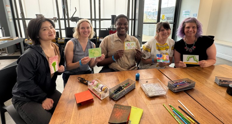 five people sitting at a table in a bright room each holding up a handmade piece of art art supplies and notebooks are spread across the table