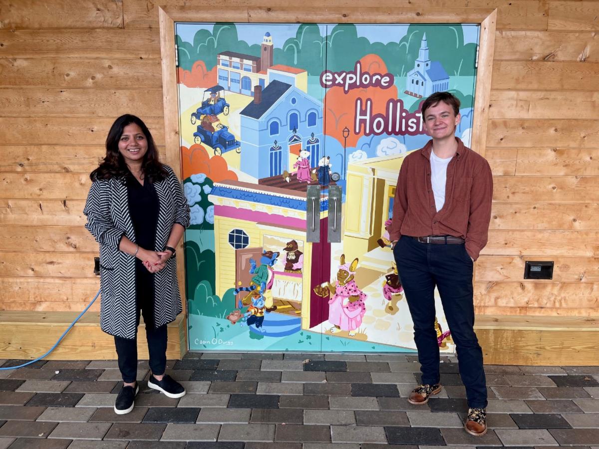 Archana Menon, former Arts and Culture Fellow, stands to the left of a colorful mural featuring whimsical animal characters and a stylized townscape with the words "Explore Holliston." The mural artist stands to the right. The mural is part of Menon's project, the Regional Trails Creative Placemaking Strategy. The background consists of a wooden wall and tiled flooring.