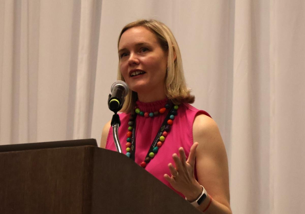 A woman stands behind a podium speaking into a microphone wearing a bright pink sleeveless top and a colorful beaded necklace with a white curtain backdrop.