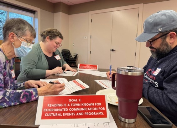 Three Reading community members are seated at a wooden table to review proposed actions for Goal 2: Reading is a town known for coordinated communication for cultural events and programs. 