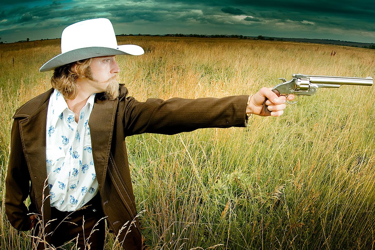 Josh Blue, dressed as cowboy, points a gun with prairie in background