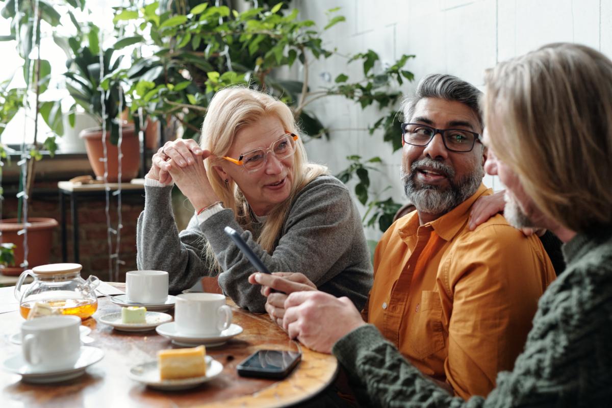 Group of mature friends gathering in cozy cafe enjoying coffee and pastries while discussing ideas. Hanging out in relaxed friendly atmosphere with plants and sunlight streaming in