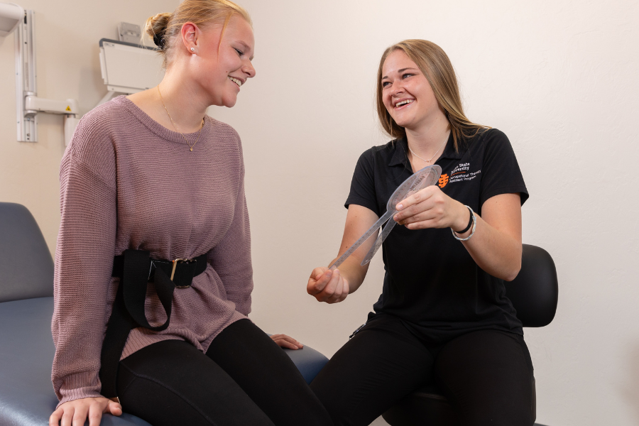 A woman sits on a doctor's table and talks to another ISU student