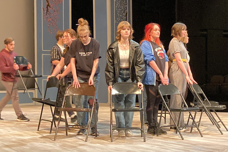 Century High School students perform on stage. They are in contemporary high school clothes and stand around a ring of chairs with dramatic/scared facial expressions.