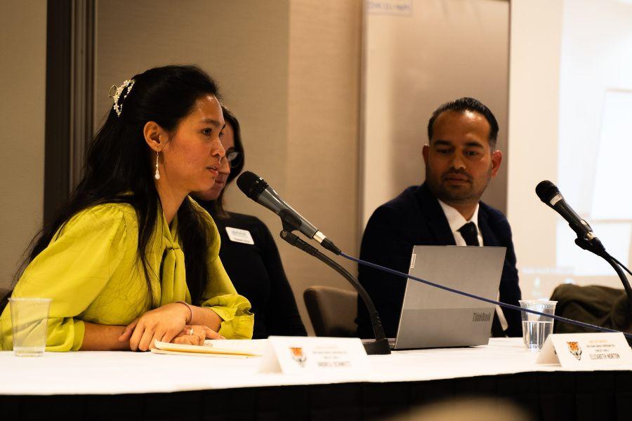 Panelists sit at a table and one of them is speaking in a microphone