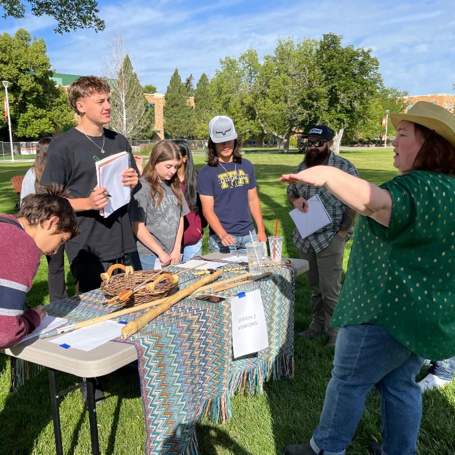 Professor Liz Redd gives an overview of the prehistoric weapons on display. She stands with students around a table set up outside on the ISU quad. The table displays replicas of prehistoric weapons made mostly from wook.