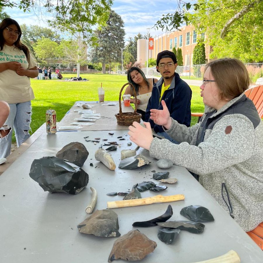 Student volunteer James Kickham demonstrates stone tool making to Discover Anthropology students. On the table are a number of pieces of rock and stone tools. Students stand or sit around the table to watch him explain.