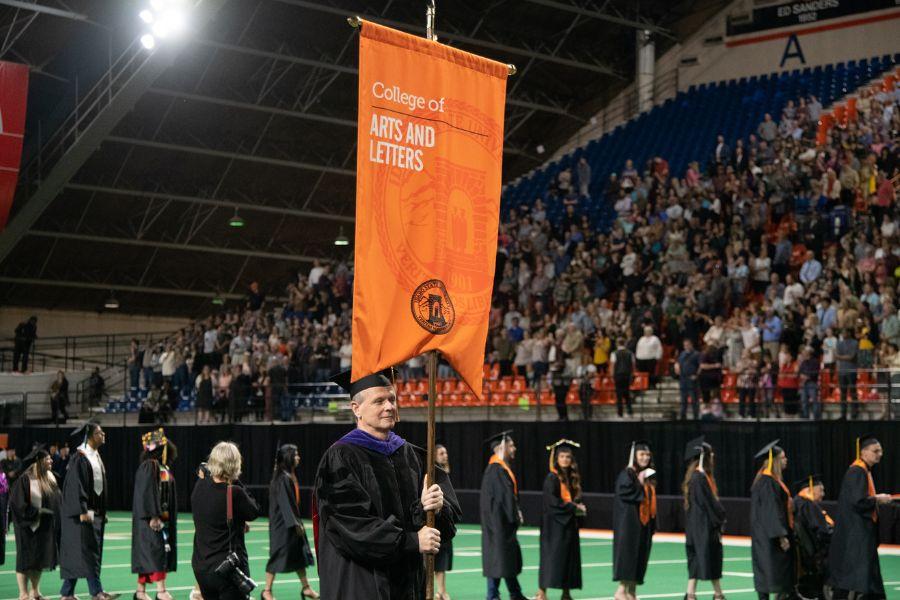 Photo of commencement in the Holt Arena. A line of students in cap and gown follow behind the flag bearer, who carries a banner that says "College of Arts and Letters". The audience can be seen in the background in the stadium seats.