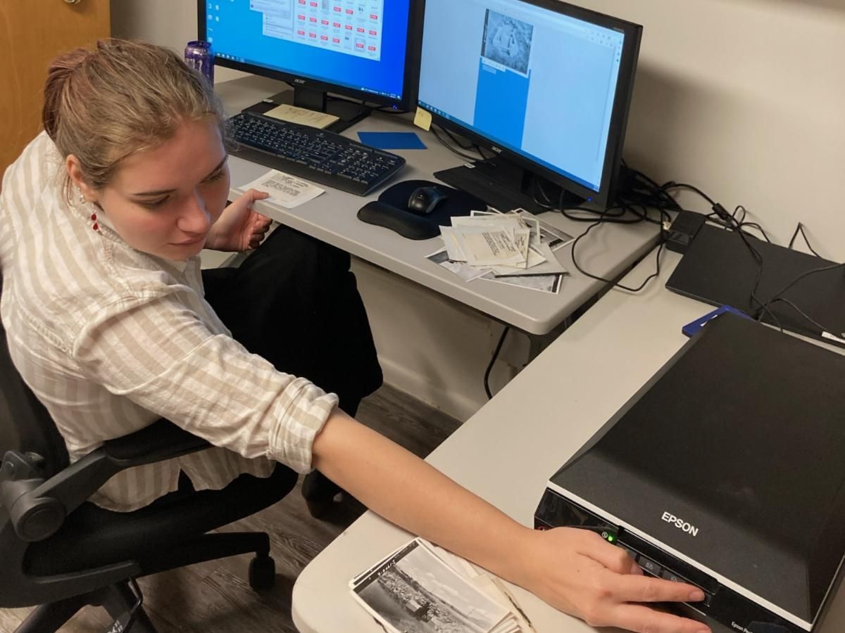 Intern student Katherine Selvage sits at her work desk with two monitors, and reaches toward a scanning machine