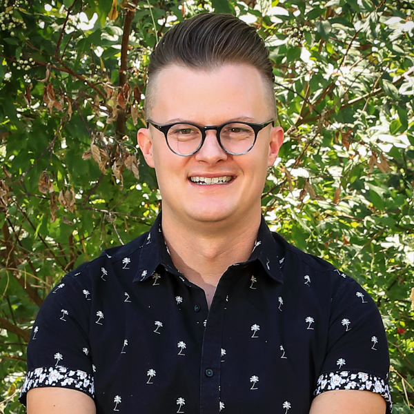 Headshot of Christopher Thomas. He sits outside with a background of green tree leaves.