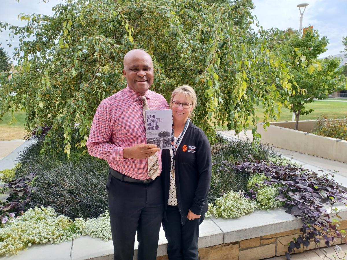 Raphael and Kandi stand in front of a garden bed with a tree in the background. Raphael holds a copy of his book