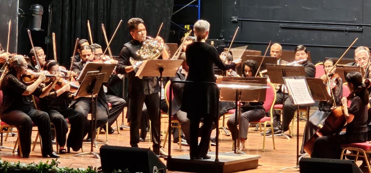 Nell Flanders stands on a stage in front of the Panama National symphony orchestra with guest artist Alberto Lin playing French horn and standing next to her