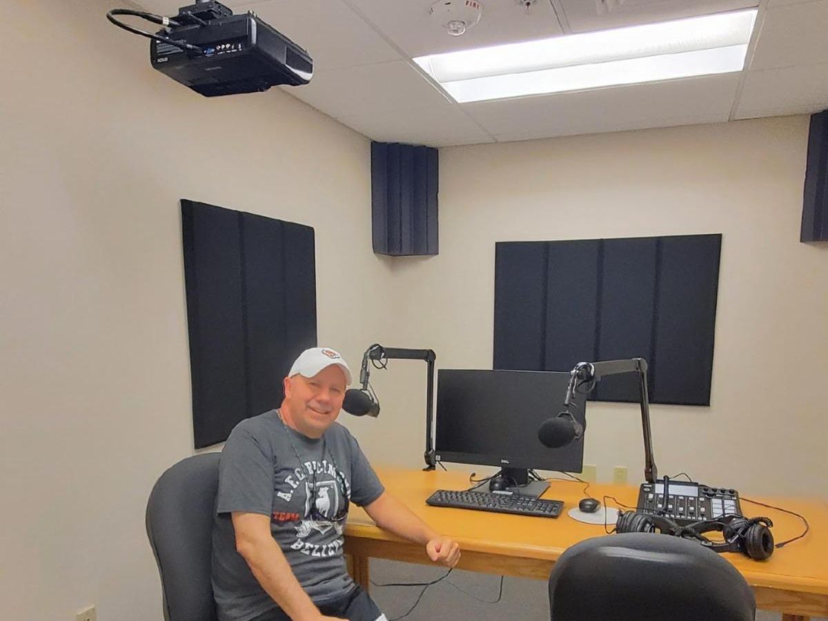 Jim DiSanza sits in one of the office chairs at the podcast studio desk. The room is equipped with audio recording equipment.