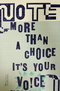 an ink print with a checkered background and text stamped in dark blue says "Vote. more than a choice it's your voice"