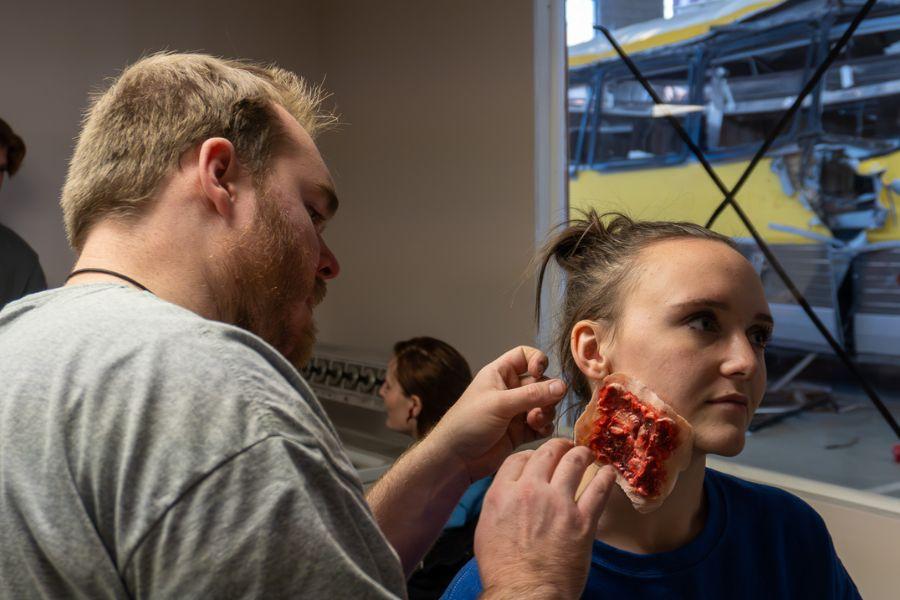 A makeup artist adds makeup to a volunteer to simulate a neck injury with flesh exposed