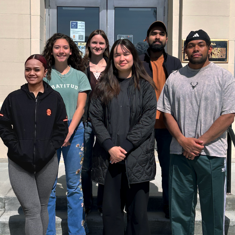 six students stand together in front of a building for a group photo
