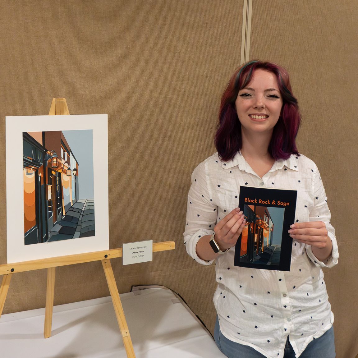 Black Rock and Sage cover artist stands next to the original art piece, a collage of a building and street. She holds the journal in front of her, which is the cover with her original artwork on it.
