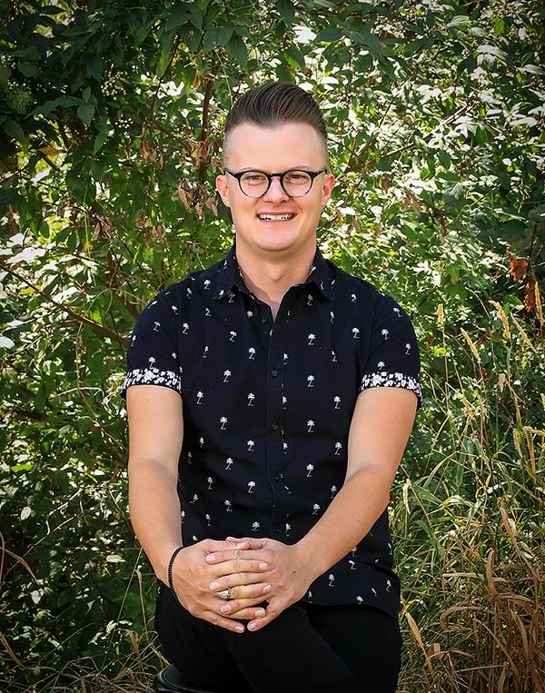 Headshot of Christopher Thomas. He is sitting in front of green foliage