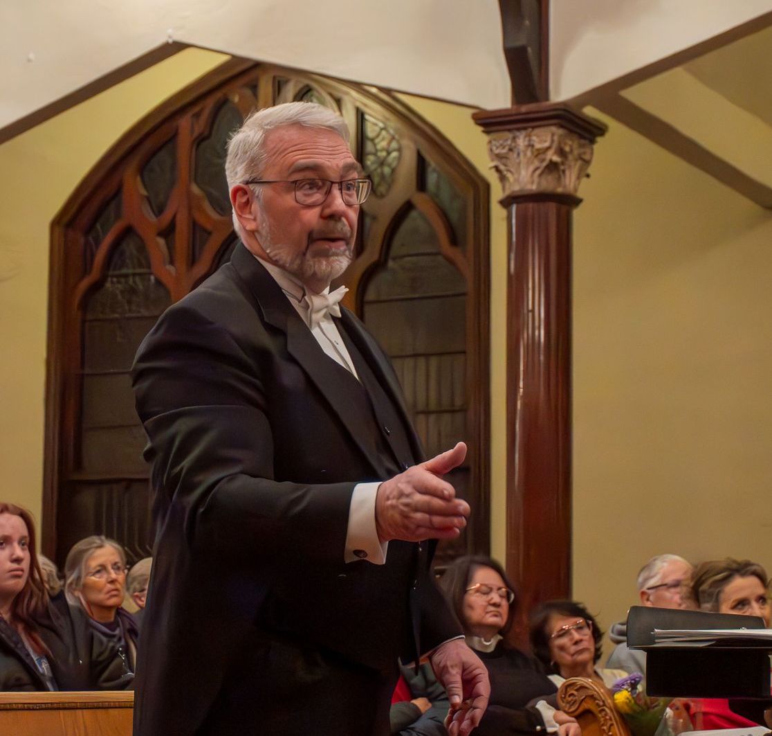 Dr. Anderson is wearing a tux and conducting a choir. His arm is uplifted.