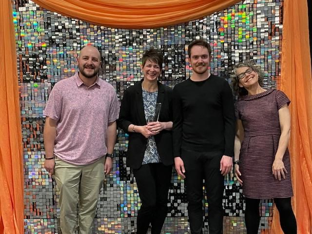 4 people stand in front of a celebratory backdrop holding plaque for winning award