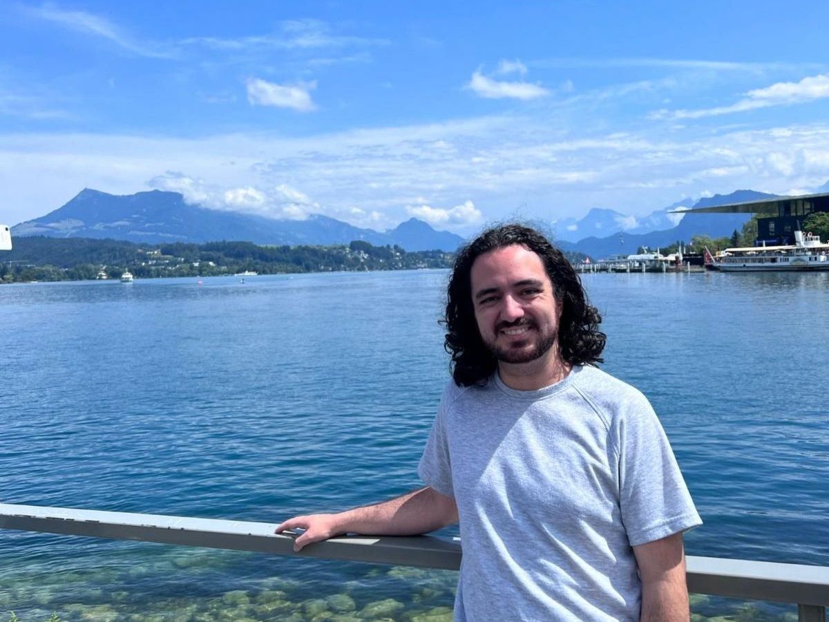 Gabe Lowman stands in front of a body of water in Switzerland with mountains and boats in the background. The sky and water are both very blue. It is daytime.