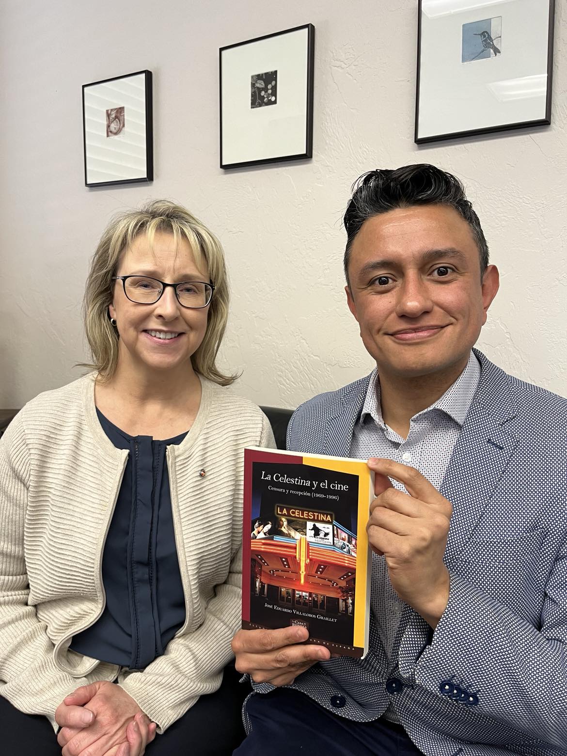 Kandi and Eduardo sit together on a couch and pose for a picture. He is holding his recently published book.