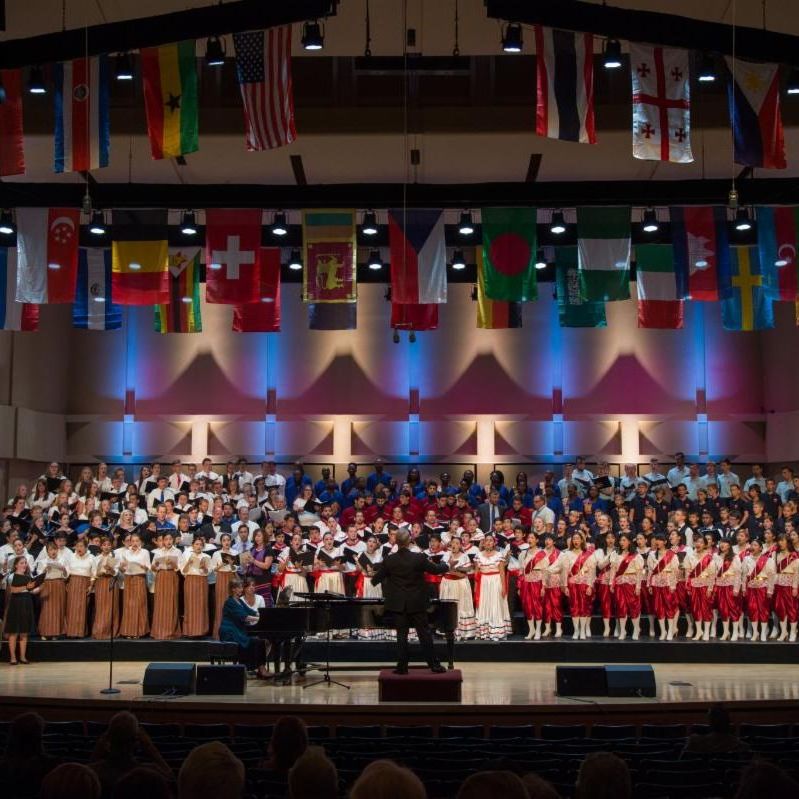 The 2019 massed choirs concert. The stage is full of choirs and is decorated from the ceiling with flags representing numerous countries.