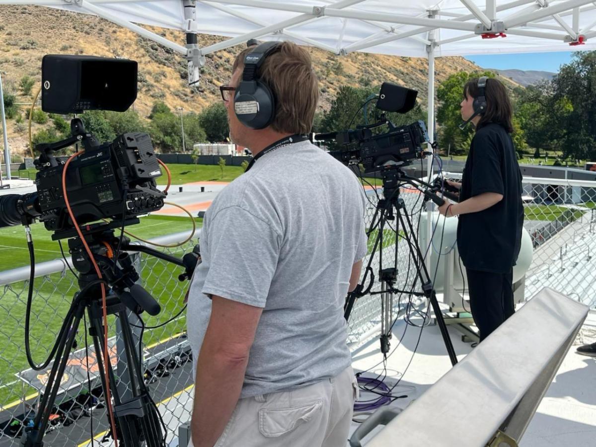 Members of the ISU sports film crew film the 1st women's soccer game of the season Fall 2023. One student stands behind a camera pointed toward the soccer field.