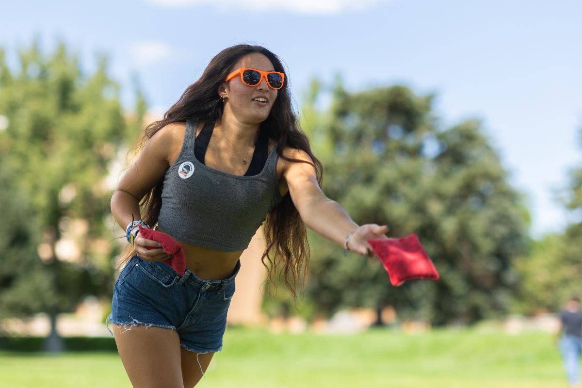 An ISU student is in the middle of throwing a bean bag during a cornhole competition. She is wearing bright orange sunglasses.