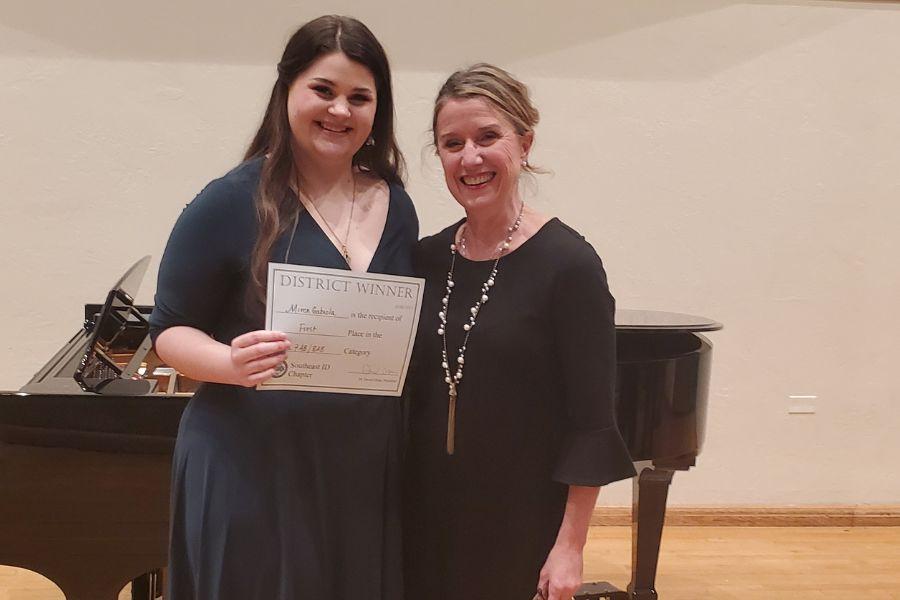 Miren Gabiola and Diana Livingston Friedley pose in front of a grand piano. Miren holds an award certificate.