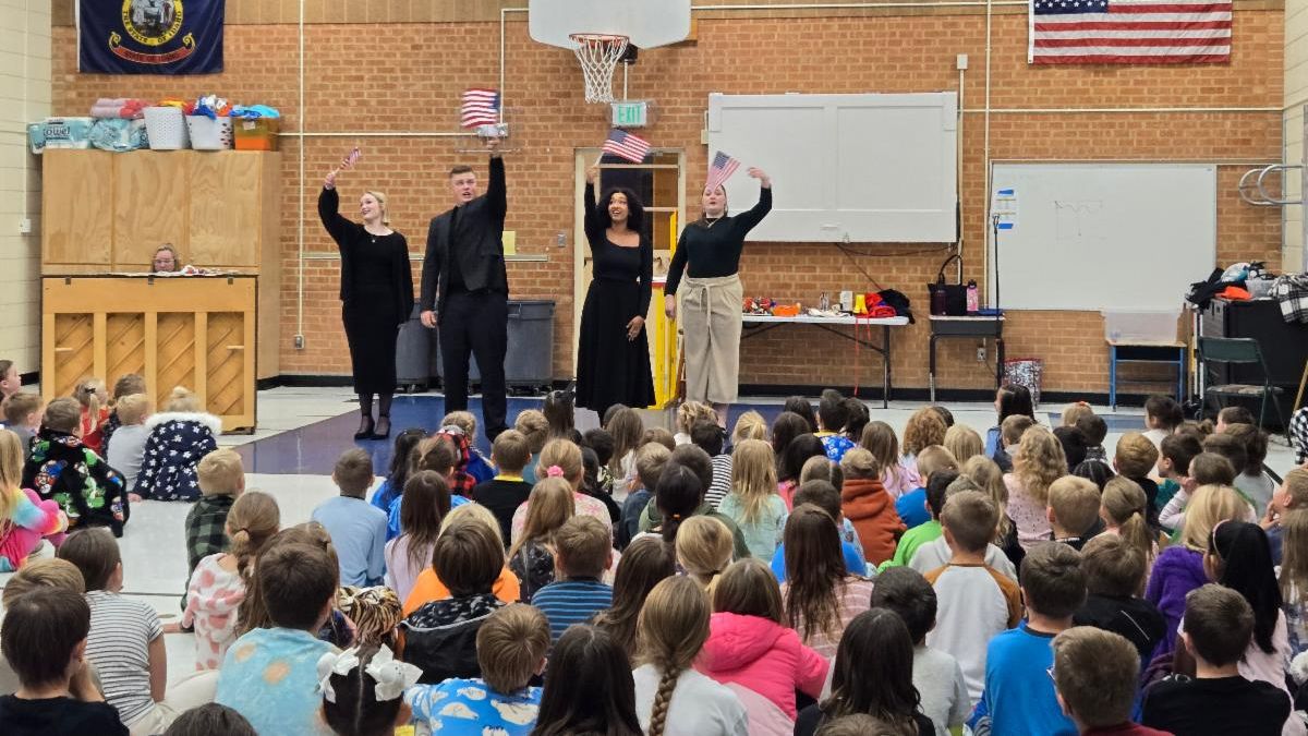 Performers waving American flags entertain children in a school gymnasium.