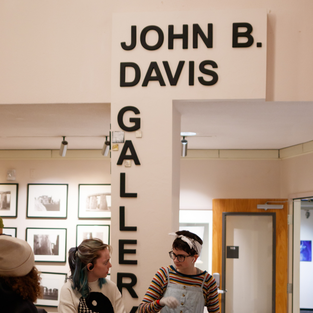 The entrance to the John B. Davis Gallery has big, black block letters naming it. Two girls stand outside the door talking. There are pictures on the wall behind.