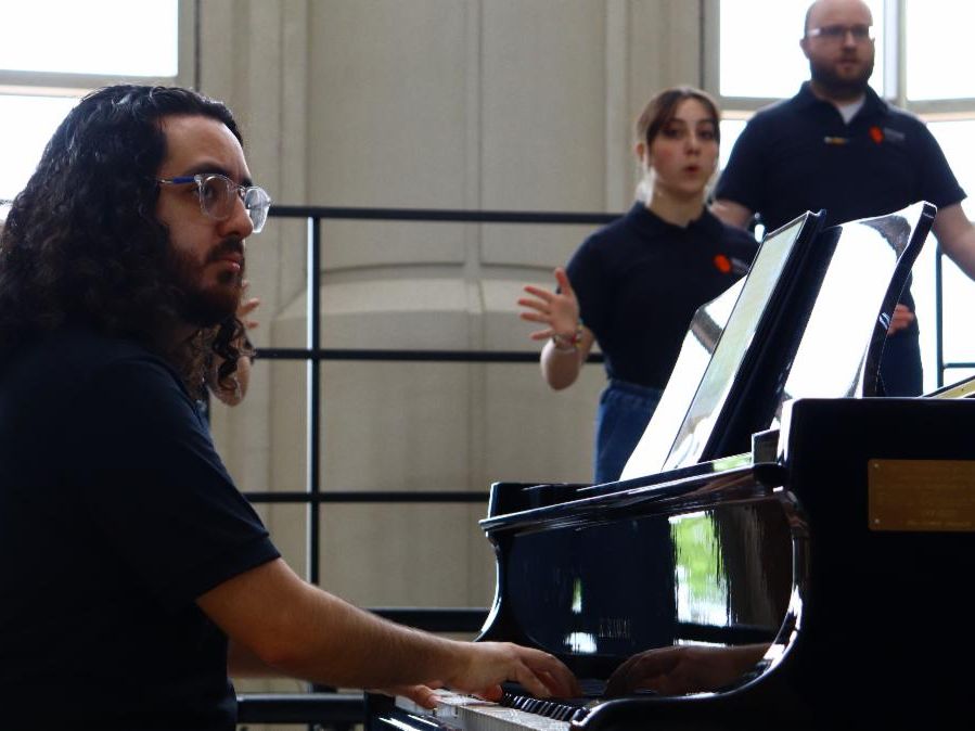 Gabe Lowman sits at a black grand piano looking toward the director. He is playing piano with the choir behind him standing on risers.