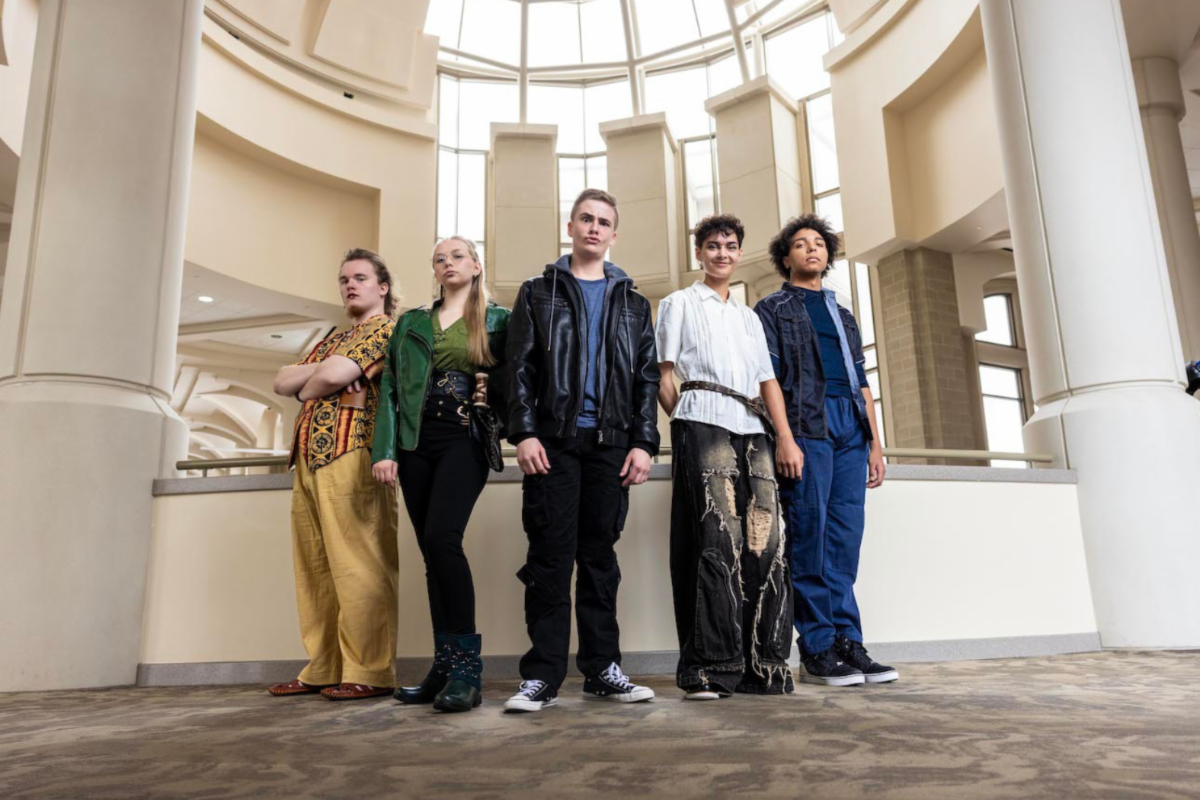  5 members of the cast (the main characters) stand in the rotunda in front of pillars. They have serious facial expressions.