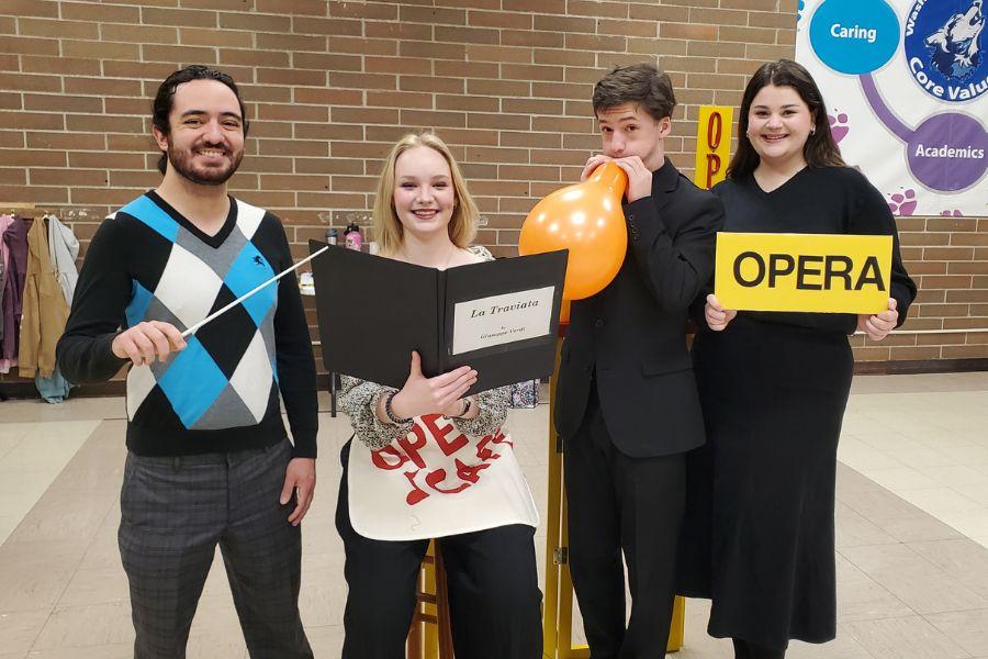 ISU students pose for a picture: one holds a music baton, one holds a music score, one is blowing up an orange balloon, and one is holding a sign that says "opera"