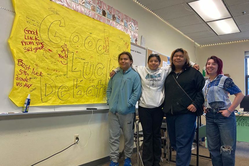 Luzahan Matsaw, Aiyana Eschief and Seth Stacey stand next to their debate coach, Abbey Vaughn, and a poster wishing them luck at the National Speech & Debate Tournament