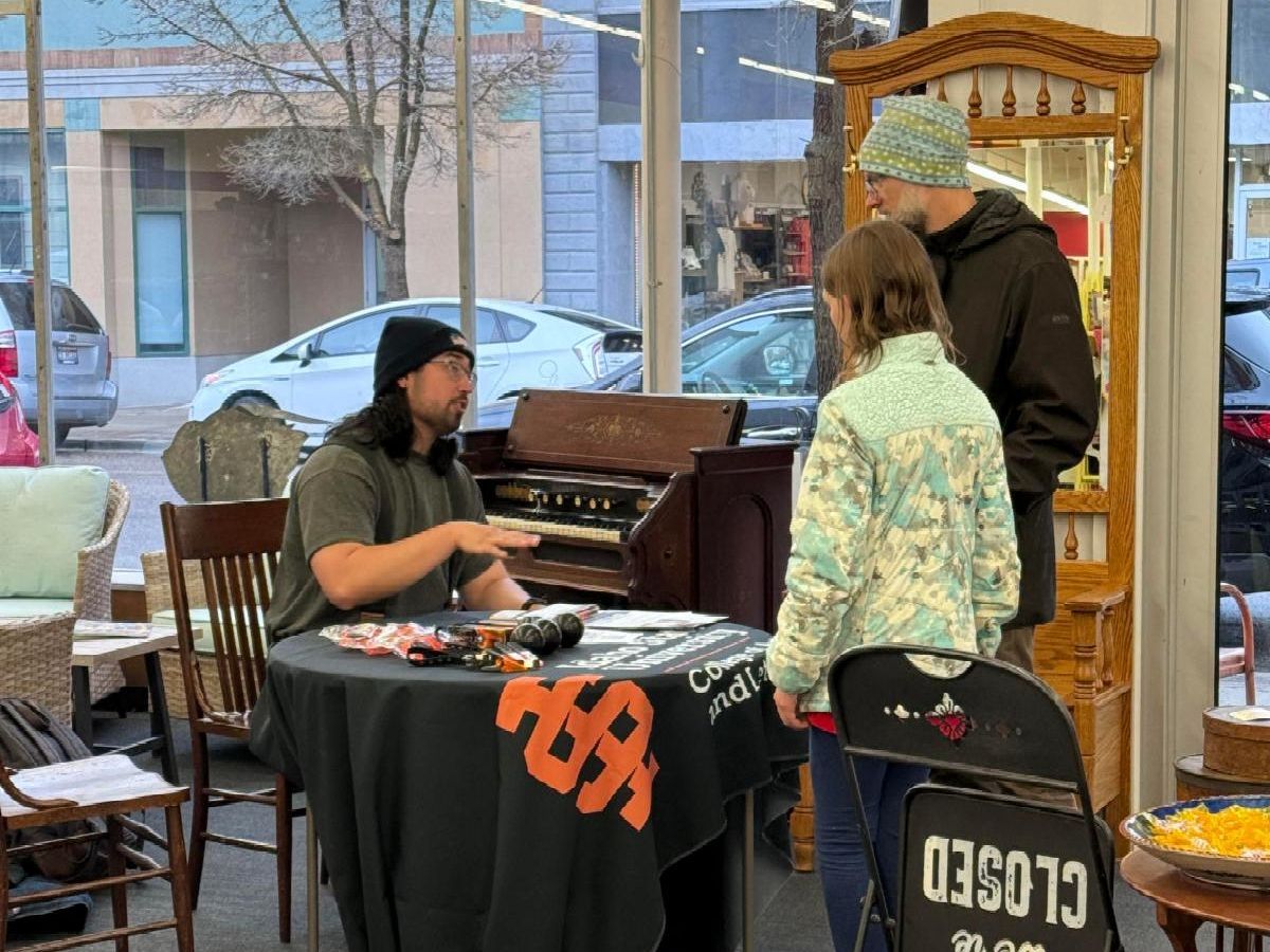 Dean Clinton sits at a table and talks to two people who are standing next to the table in the antique shop