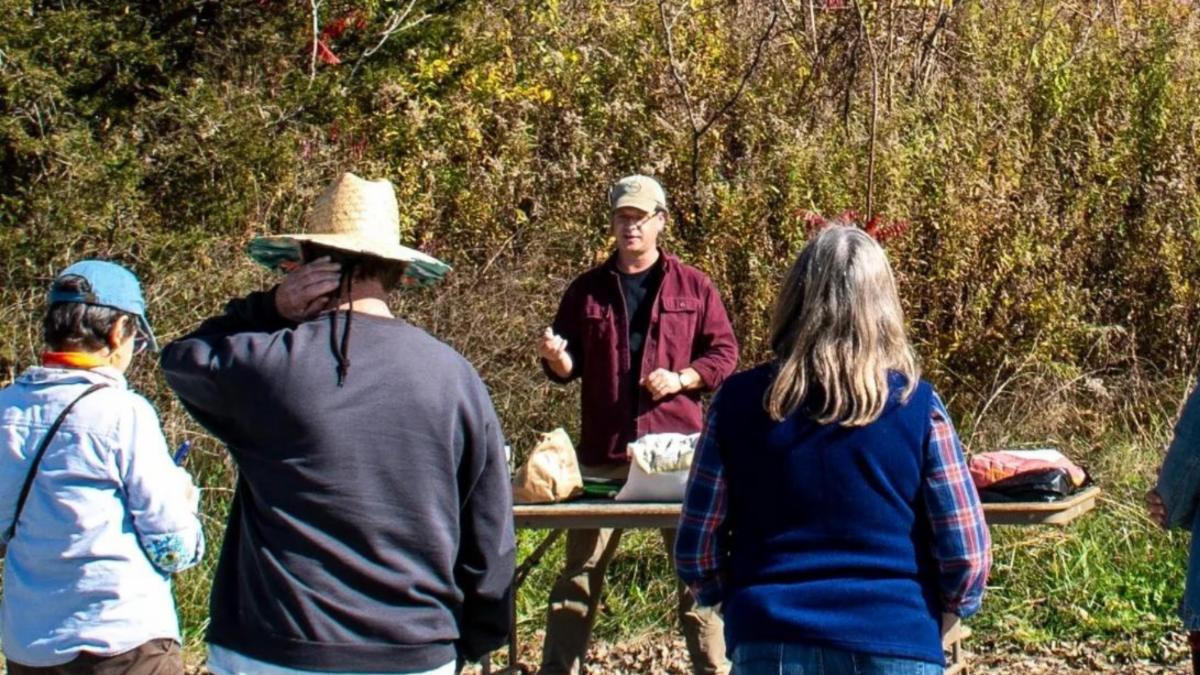 Brian Collier leads a hands-on workshop. He is pictured outdoors at a picnic table with a small crowd gathered around listening to him.