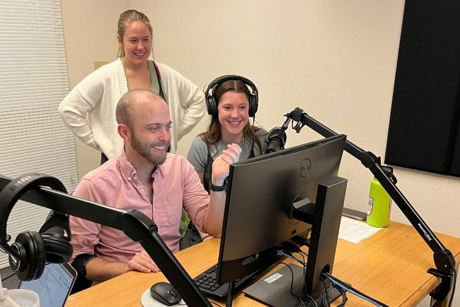Daniel Shelden, Labelle Knittel, and Saybree Swanson stand and sit at a desk, smiling, with recording equipment