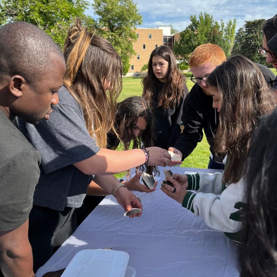 A group of students gather around a table on the ISU quad, piecing together parts of a broken piece of pottery.