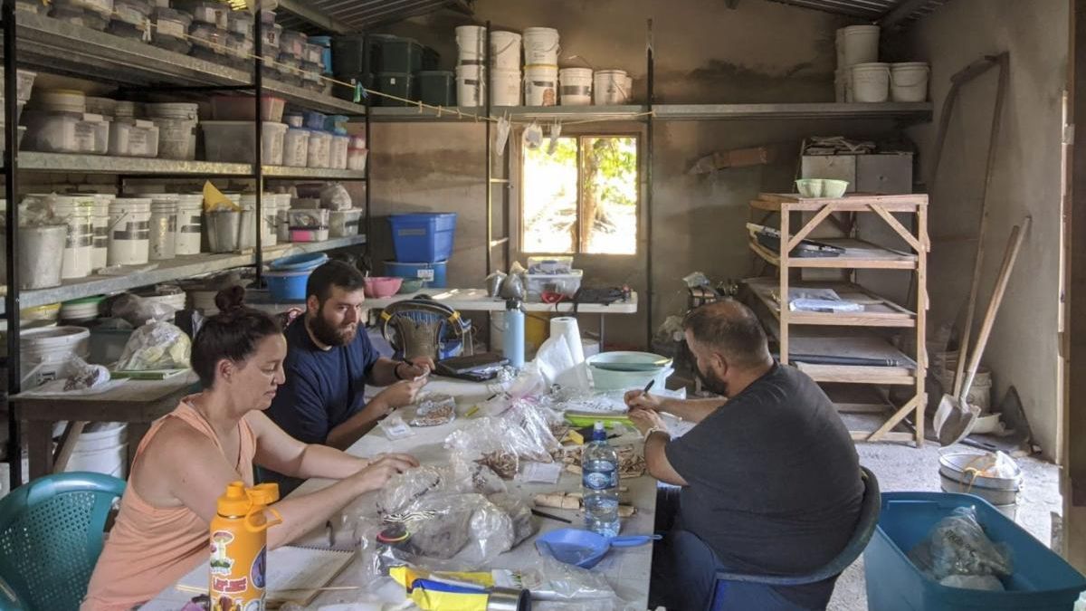 Three people working at a cluttered table in a workshop with shelves of containers.