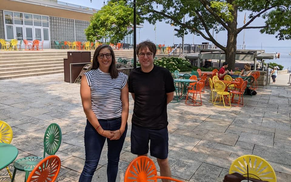 Laufenberg and Martinez stand outside in the daytime on a stone-paved area with colorful chairs and tables and a tree in the background