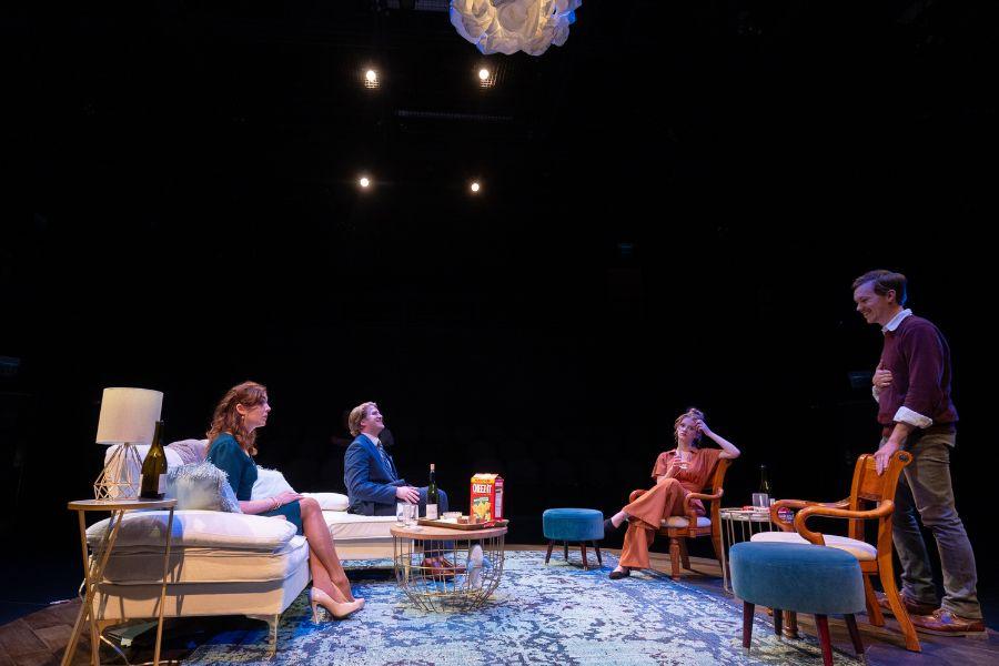 A scene of a performance of Life X 3. 4 actors are on the stage. The stage is set up as a home with chairs and tables. 