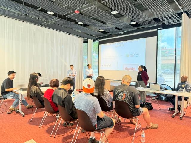 A community meeting or presentation takes place in a modern conference room with floor-to-ceiling windows, white curtains, and a large projection screen. Two presenters stand at the front of the room addressing approximately a dozen attendees seated in rows of chairs. Participants of various ages are listening attentively, some taking notes or looking toward the speakers. Additional facilitators are seated at tables on either side of the room. The space features exposed industrial-style ceilings, red carpet flooring, and abundant natural light streaming through the windows. A presentation slide is displayed on the screen behind the speakers.