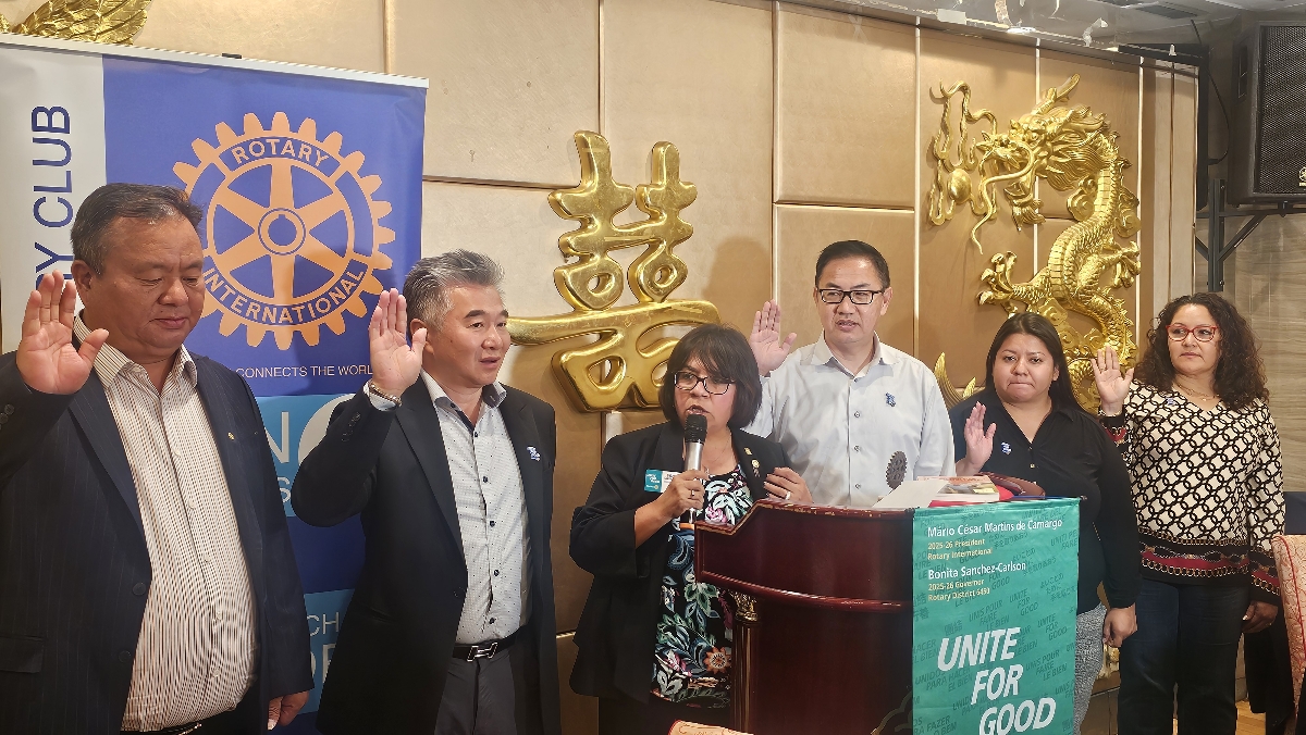 A group of six people stands with raised right hands some reciting an oath at a podium with Rotary Club and Unite for Good banners in the background