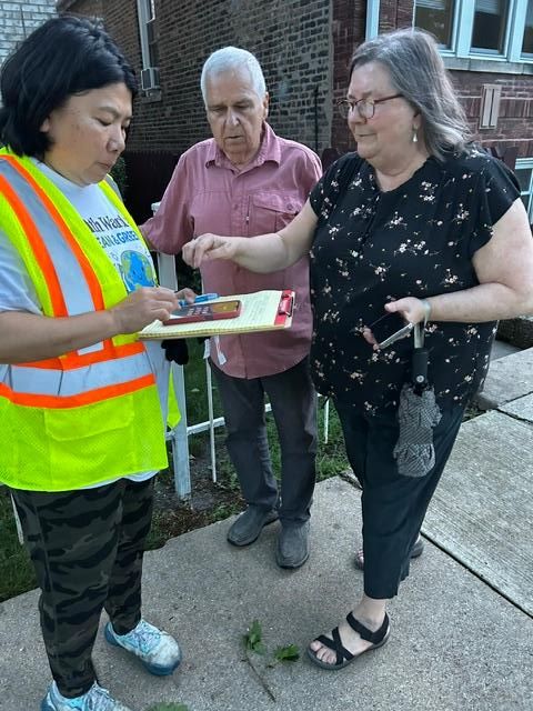 A person wearing a high-visibility safety vest stands on a residential sidewalk speaking with two older residents outside a brick apartment building. One resident points to a clipboard while the person in the safety vest takes notes or reviews information on a smartphone. The other resident stands nearby listening. The interaction appears to be part of a neighborhood outreach effort, community assessment, or response to a local issue. The scene takes place on a tree-lined residential street with multi-unit brick homes in the background.